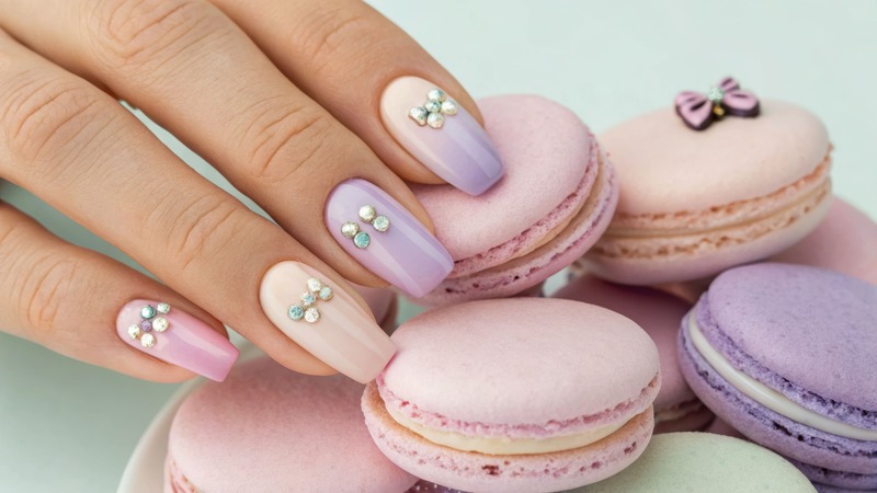 Macaron nails; mano con smalto color pastello e decorazione di fiocco; le dita si poggiano su macaron colorati