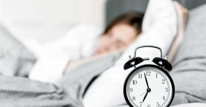 Quando il rumore bianco aiuta davvero a dormire e concentrarsi Quando il rumore bianco aiuta davvero a dormire e concentrarsi: Una sveglia retro si trova accanto a una donna che dorme in…